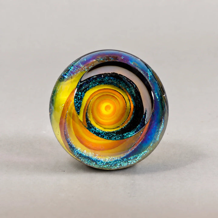 A round glass paperweight featuring a vibrant yellow and orange spiral pattern, accented by a dark blue glitter ring and iridescent rainbow reflections on a neutral gray background.