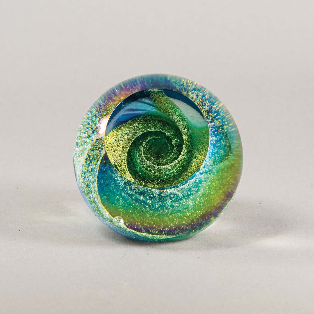 A round glass paperweight featuring a vibrant green and blue spiral design with shimmering gold glitter and iridescent purple-blue edges, set against a neutral light gray background.