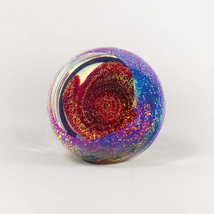 A round glass paperweight featuring a vibrant, swirling design with a red and gold spiral center, surrounded by purple and blue outer swirls with embedded glitter, set against a plain light gray background.