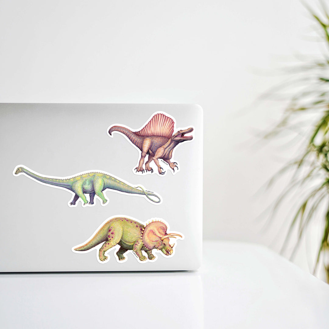 Three dinosaur stickers on a laptop with a blurred plant in the background
