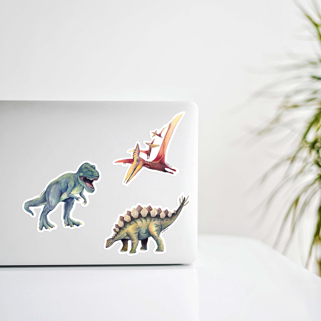 Three dinosaur stickers on a laptop with a blurred plant in the background