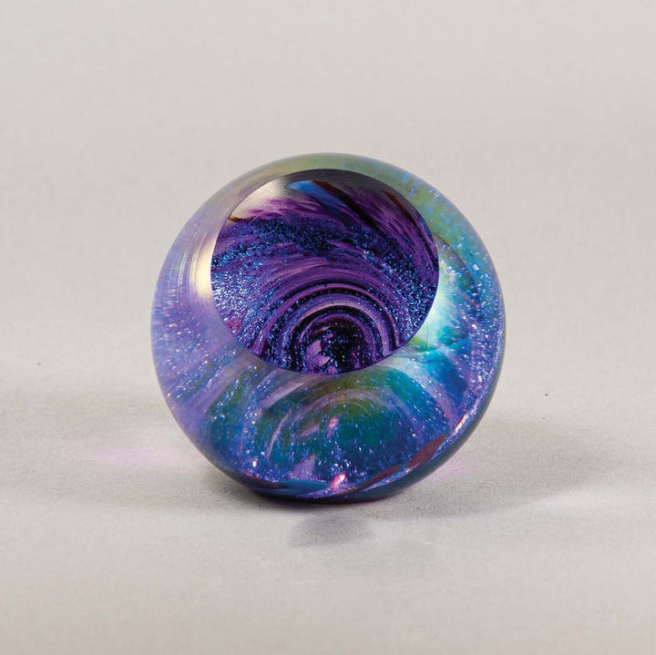 A round glass paperweight with a swirling galaxy pattern in purple, blue, and green, set against a neutral light gray background.