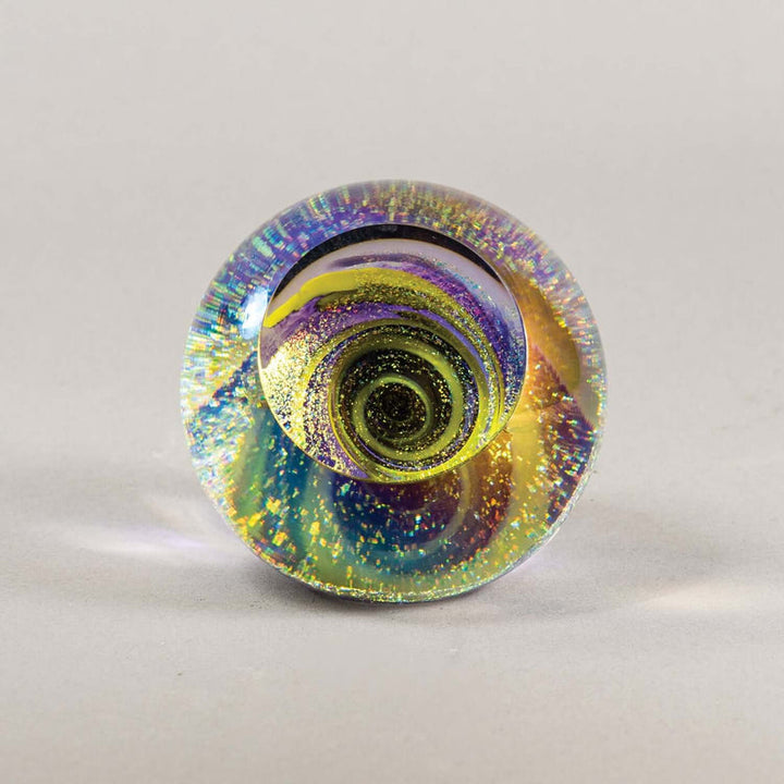A round glass paperweight featuring a vibrant, swirling pattern of iridescent colors including purple, blue, green, and yellow, with fine glitter suspended throughout, set against a neutral light gray background.