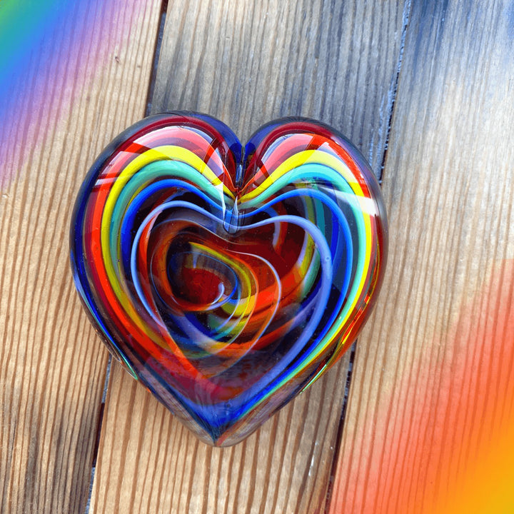 A heart-shaped glass paperweight with concentric swirls of red, orange, yellow, green, blue, and purple, resting on a rustic wooden surface.