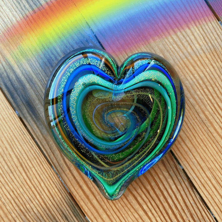 A heart-shaped glass art piece with swirling blue, green, and gold glitter patterns, resting on a wooden surface with a rainbow reflection in the background.