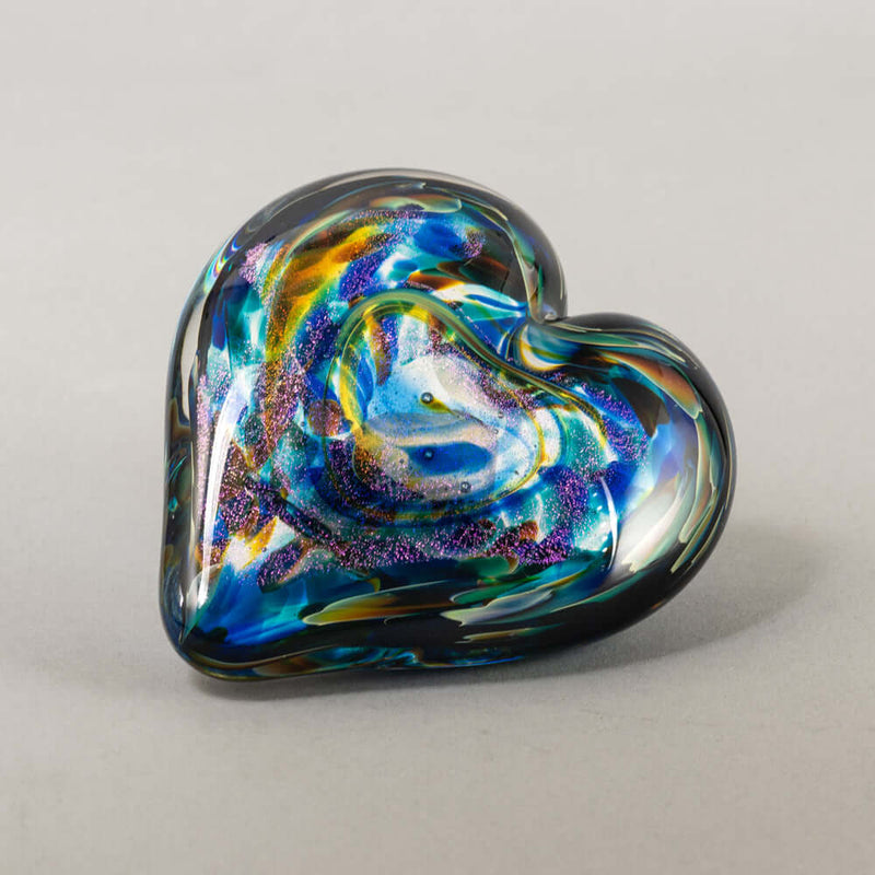 A heart-shaped handblown glass art piece with swirling iridescent colors of blue, green, purple, and gold on a light gray background.