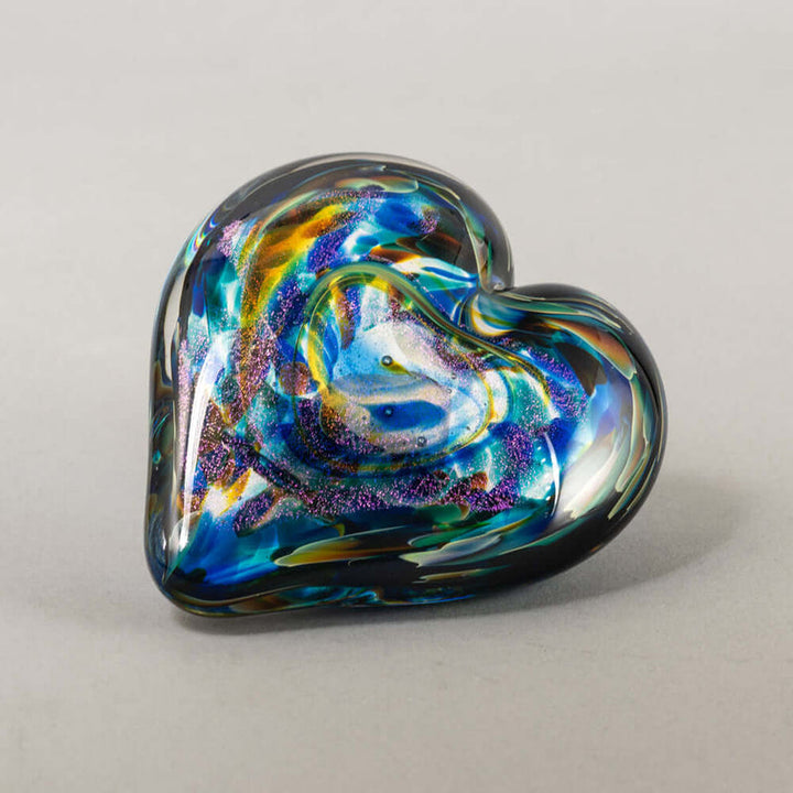 A heart-shaped handblown glass art piece with swirling iridescent colors of blue, green, purple, and gold on a light gray background.