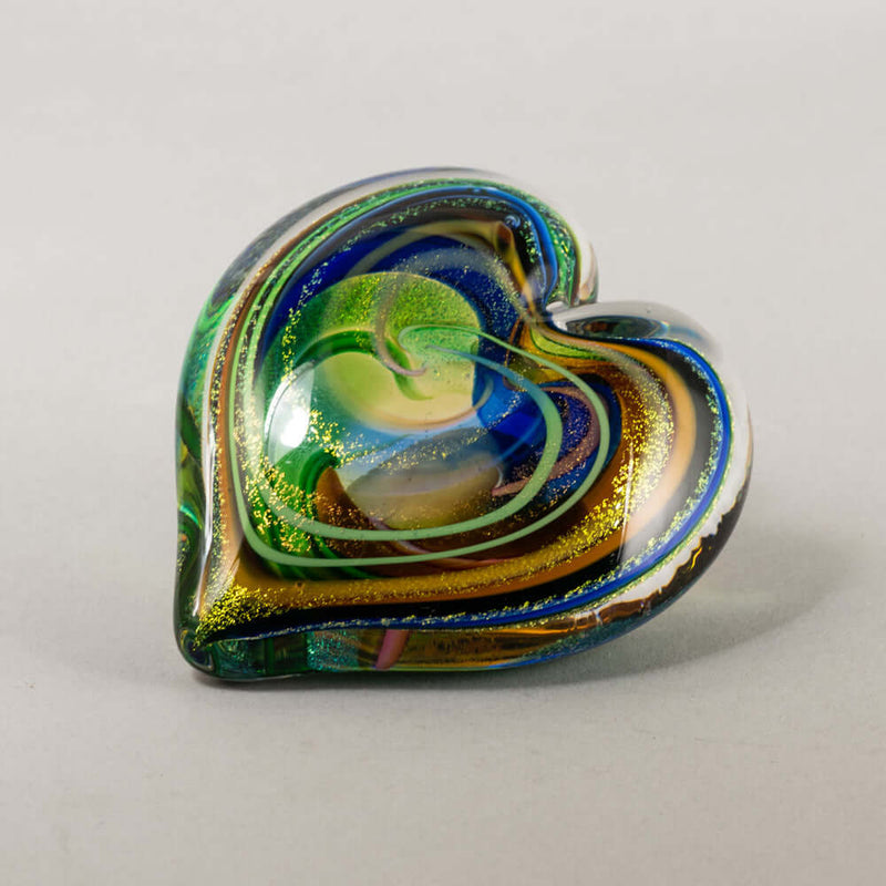 A heart-shaped glass art piece with swirling layers of blue, green, gold, and brown on a light gray background.