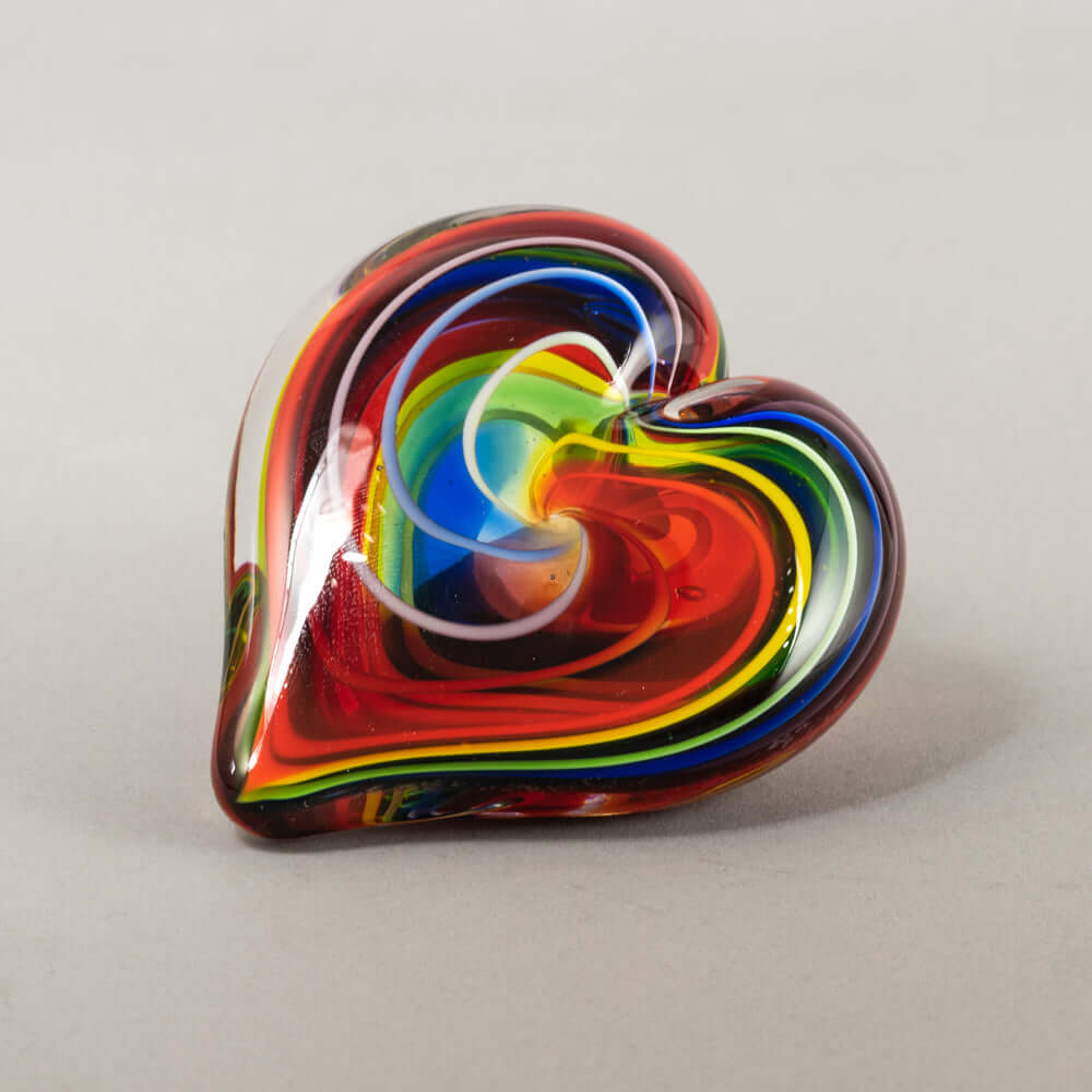 A heart-shaped glass figurine with a vibrant swirl pattern of red, orange, yellow, green, blue, and white, photographed on a neutral light gray background.