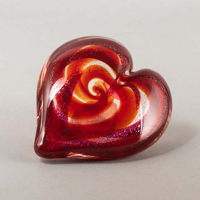 A heart-shaped glass sculpture featuring a swirling rose pattern in vibrant red and orange with fine silver glitter, set against a neutral light gray background.