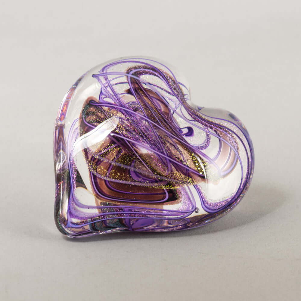 A heart-shaped clear glass paperweight featuring swirls of purple, white, and metallic gold glitter, photographed on a neutral light gray background.