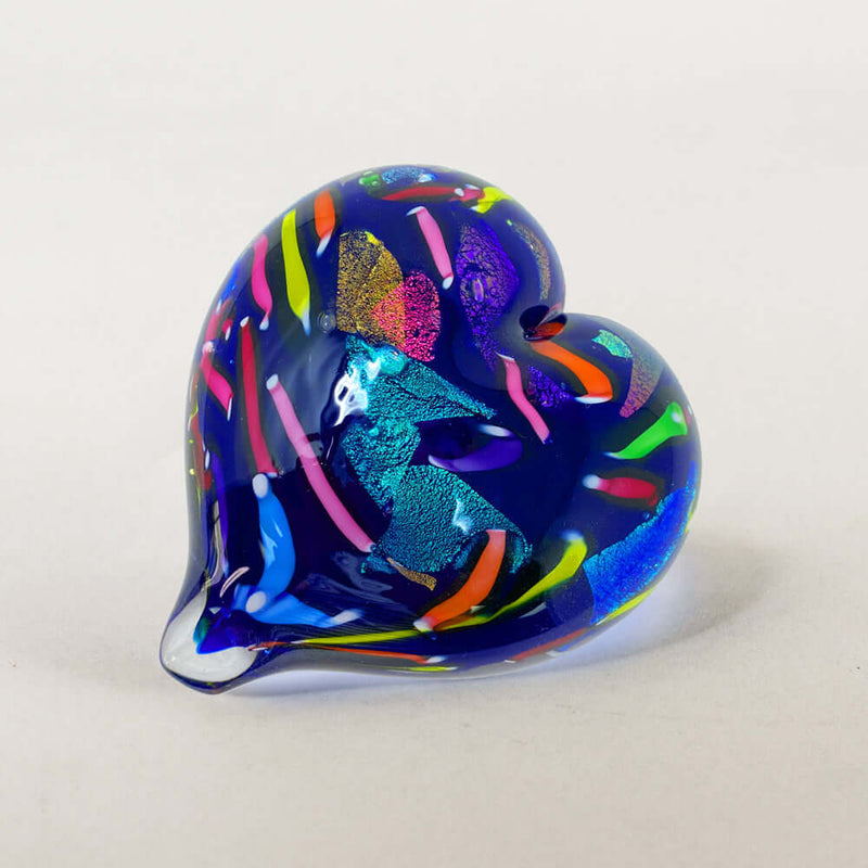 A handcrafted glass heart figurine with a deep blue base and vibrant, multicolored swirls and stripes, photographed on a neutral light gray background.