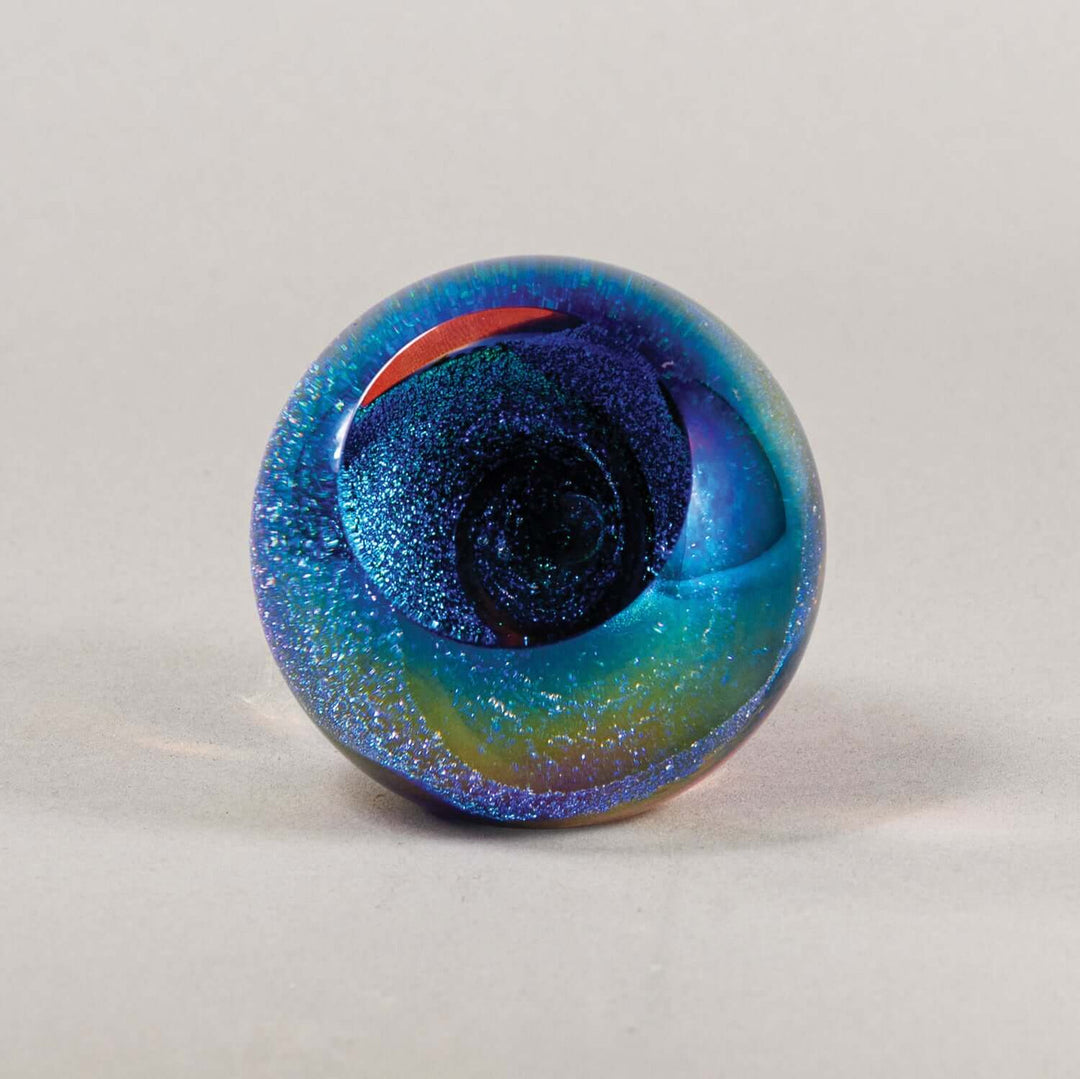A round glass paperweight featuring a swirling galaxy design with blue, teal, and purple hues, embedded with fine glitter, and a small red accent near the top, set against a neutral light gray background.