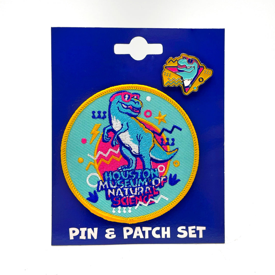 An iron-on patch and enamel pin set on  dark blue cardstock, featuring a cyan T. rex with sunglasses and the museum name
