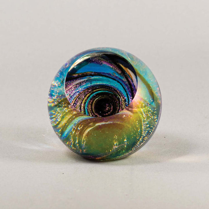 A round glass paperweight featuring a swirling marbled pattern of teal, green, gold, purple, and black, photographed on a neutral light gray background.