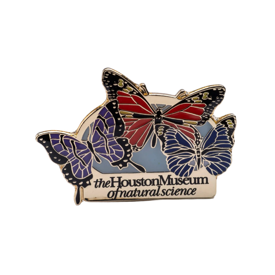 An enamel pin with a multicolored butterfly design, featuring red, blue, and black colors with a gold-tone border, and the Houston Museum of Natural Science logo.